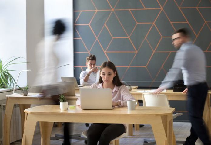Woman working diligently on a laptop in a busy, modern open-plan office. -Lenvi