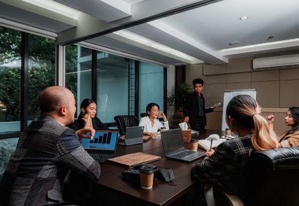 People gathered around a meeting table during a business presentation with laptops and performance data displayed on screen
