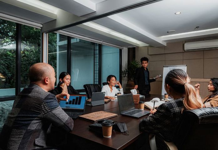 People gathered around a meeting table during a business presentation with laptops and performance data displayed on screen