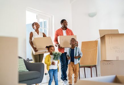 A mum and dad with their two children carry moving boxes and plants into their new home